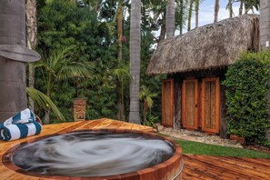 Un bain à remous en bois, entouré d’une végétation luxuriante, une cabane au toit de chaume et un banc en bois avec une serviette.