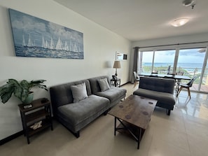 A modern living room with a sofa, a coffee table, a painting of sailboats, a potted plant, and a view of the ocean through large windows.