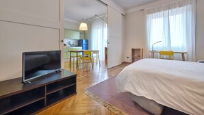 Une chambre d’hôtel avec un lit, un téléviseur posé sur un meuble, un bureau avec une chaise et un coin cuisine visible à travers une porte ouverte.