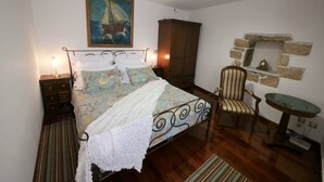 Een slaapkamer met een bed, een stoel, een nachtkastje, een dressoir, een schilderij en een plank aan een stenen muur.