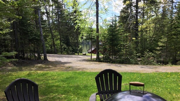 Een camping met twee Adirondack-stoelen en een metalen barbecue op de voorgrond, omgeven door een bebost gebied met een onverhard pad dat naar een hutje in de verte leidt.