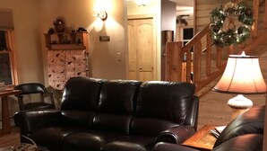 Een woonkamer met een leren hoekbank, een houten salontafel en een lamp. Er is een trap met een houten leuning en een krans aan de muur.
