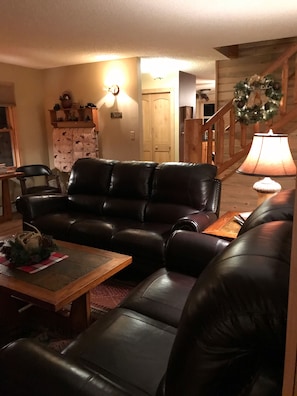 Ein Wohnzimmer mit einem ledernen Ecksofa, einem Holztisch und einer Lampe. Eine Treppe mit Holzgeländer und einem Kranz an der Wand ist ebenfalls zu sehen.