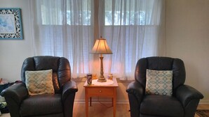 Una habitación con dos sillones de cuero negro con cojines estampados, una mesita de madera con una lámpara y objetos decorativos, y cortinas blancas traslúcidas.
