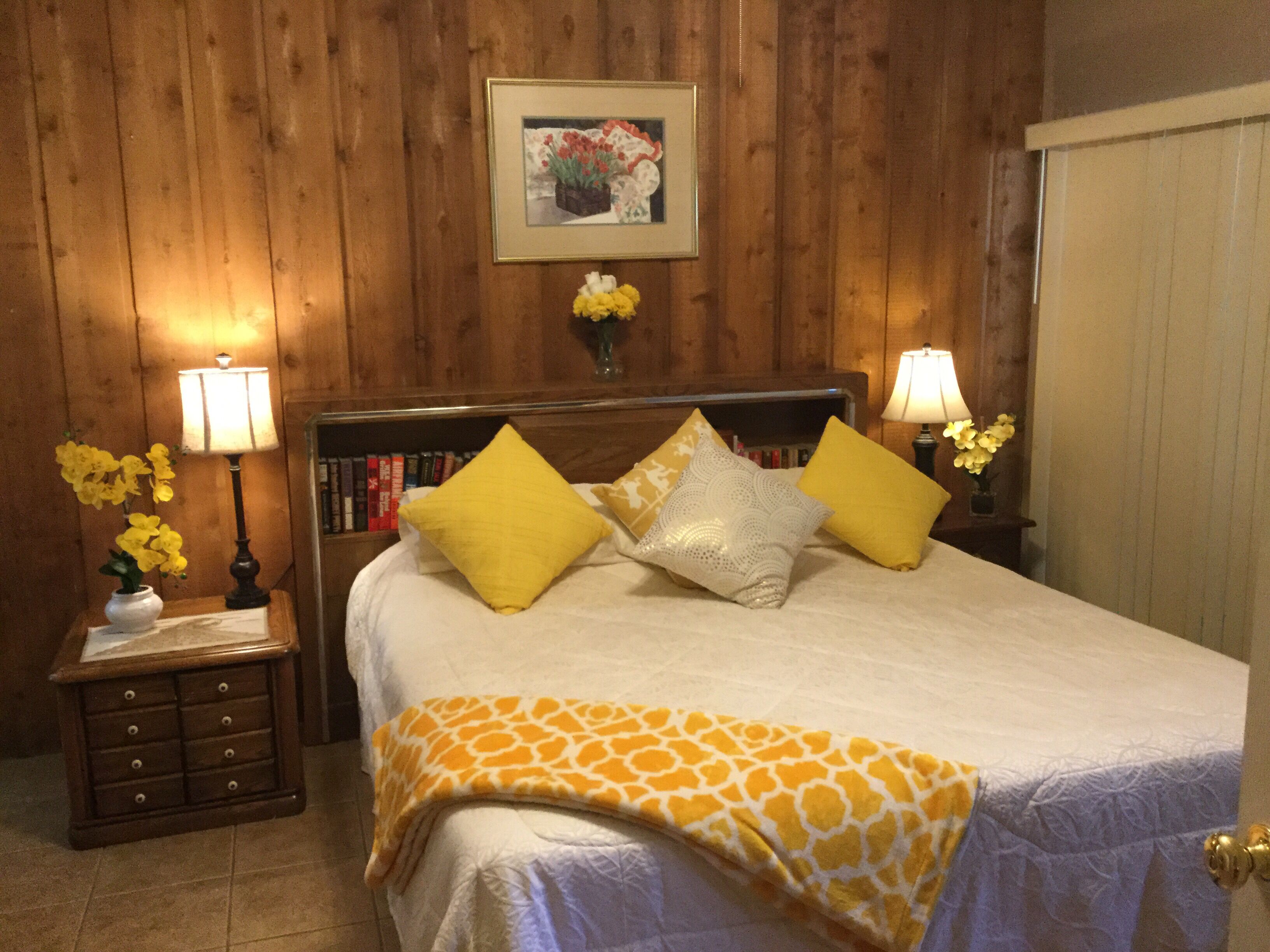 Een slaapkamer met een houten hoofdbord, een bed met een deken met een wit en geel patroon, twee gele kussens en twee bedlampen.