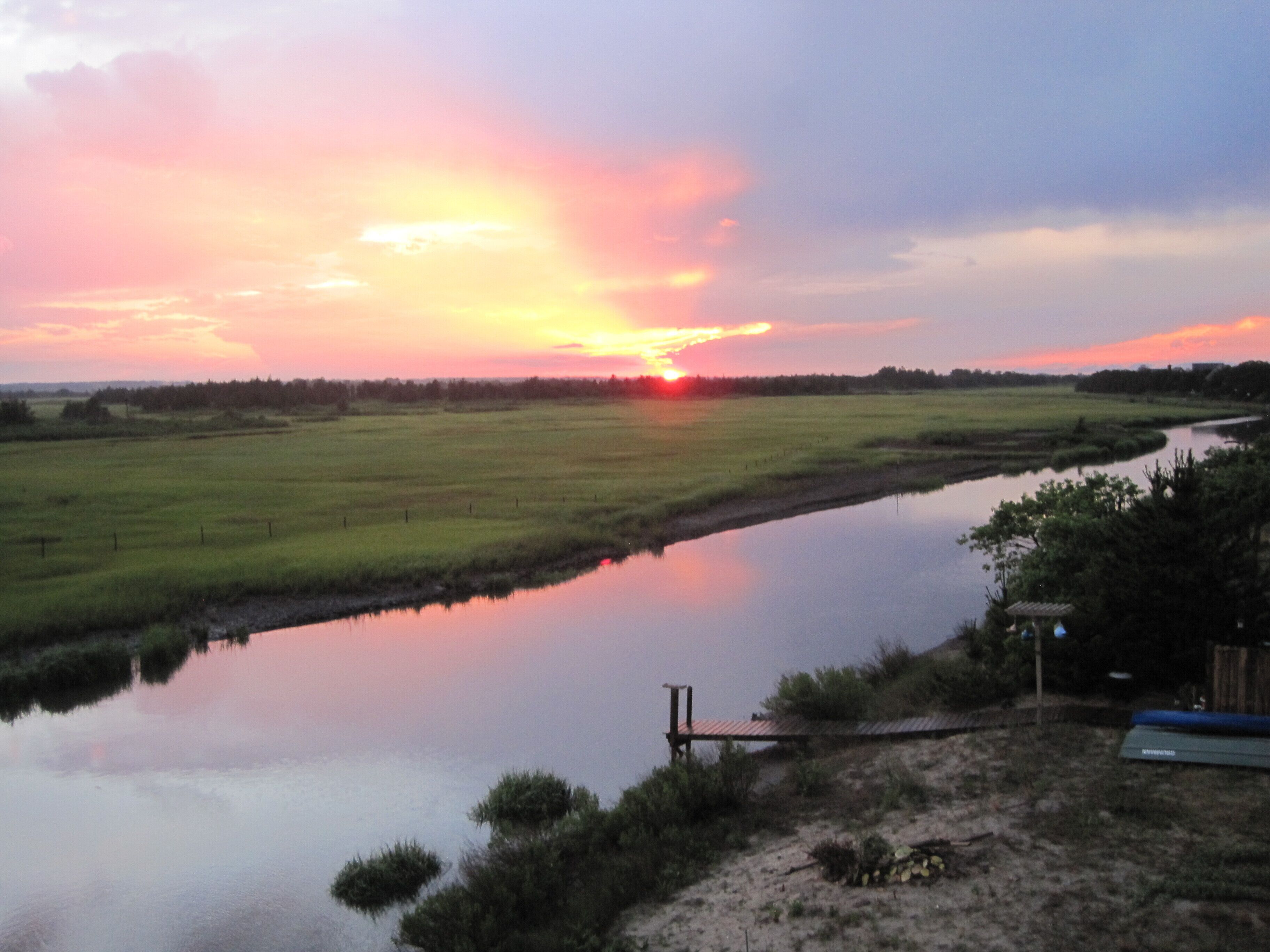 A river with a wooden dock, surrounded by grassy fields and trees.