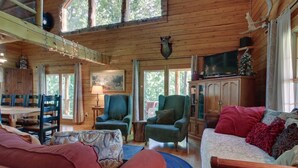 A cozy cabin interior with a dining area, two green armchairs, a sofa, and a television. - Private log cabin with hot tub and mountain views on 14 acres, pet friendly! (N.Ferrisburgh)