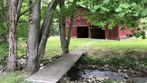 Eine rustikale Holzbrücke über einem Bach, umgeben von Bäumen und einem roten Bauernhof im Hintergrund.