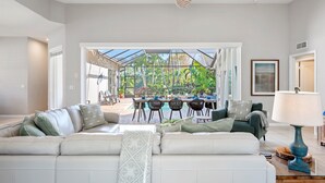 Un salón amplio con un sofá grande, una zona de comedor con mesa y sillas, y vistas a un jardín tropical a través de un ventanal.