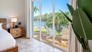 Un dormitorio con un amplio ventanal corredera que da a vistas de un lago, una mesita de noche de madera con lámpara y una cama con ropa blanca.