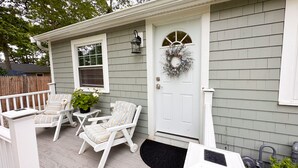 A house with a porch featuring two white chairs with striped cushions, a small table, and a potted plant. - Quaint Cozy Beach Cape Escape (East Falmouth)