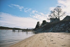 A beach with a wooden pier extending into calm waters, a sandy shore with footprints, and a house with a stone retaining wall on the right.