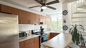 Cocina con alacenas de madera, refrigerador de acero inoxidable, microondas, estufa y ventilador de techo. Se ve una ventana con cortinas y una planta en la encimera.