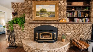Een stenen openhaard met een zwarte mantel, erboven een ingelijst schilderij en een boekenplank aan de rechterkant.
