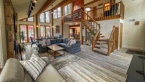 A spacious living room with a large sectional sofa, a wooden staircase, and a view of the outdoors through large windows.
