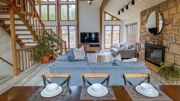 A spacious living room with a high ceiling, a staircase, a fireplace, and a dining area set for four.