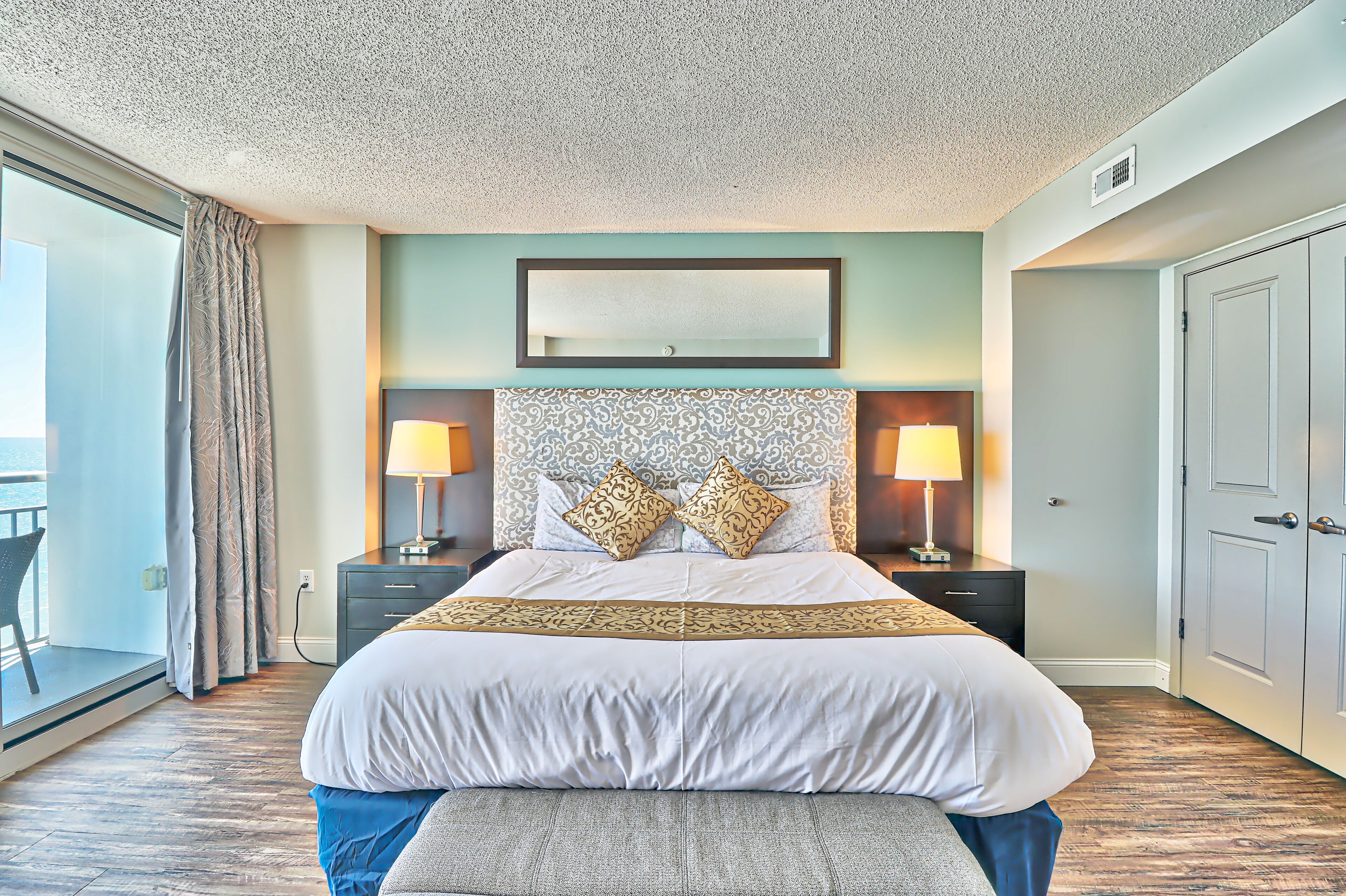 A neatly arranged bedroom with a large bed, two bedside tables with lamps, a patterned headboard, and a view to the outside.
