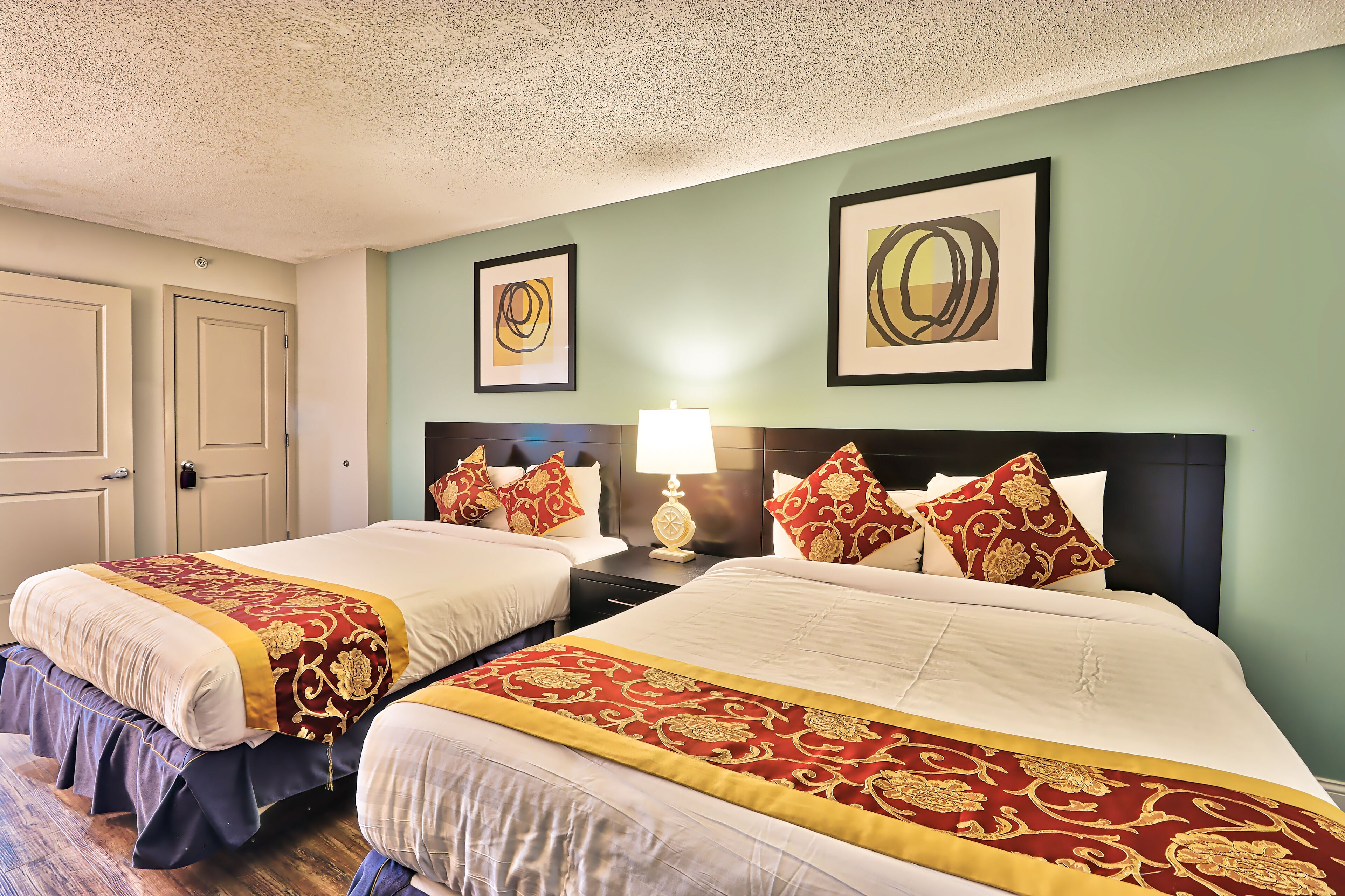 A hotel room with two beds, each with a red and gold bedspread, a bedside table with a lamp, and artwork on the walls.
