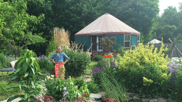Um jardim com diversas plantas e flores, uma pequena casa com telhado cônico e uma pessoa em pé no jardim.