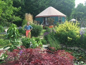 A garden with a variety of plants and flowers, a small house with a conical roof, and a person standing in the garden.