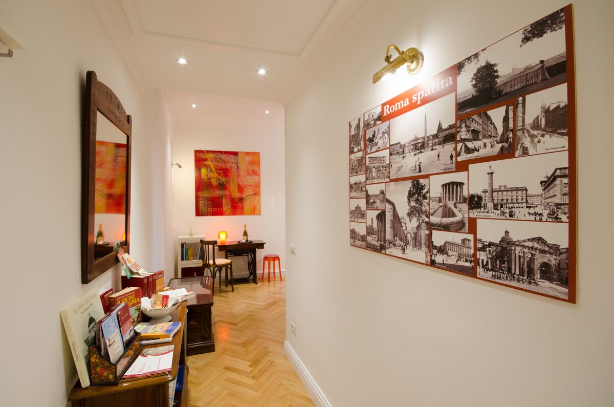 Un corridoio con una galleria di foto appesa al muro intitolata “Roma sparta”, una scrivania di legno con libri, una sedia e un piccolo tavolino con una bottiglia.