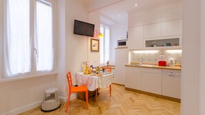 Een moderne keuken met witte kasten, een eettafel met oranje stoelen en een aan de muur bevestigde televisie.