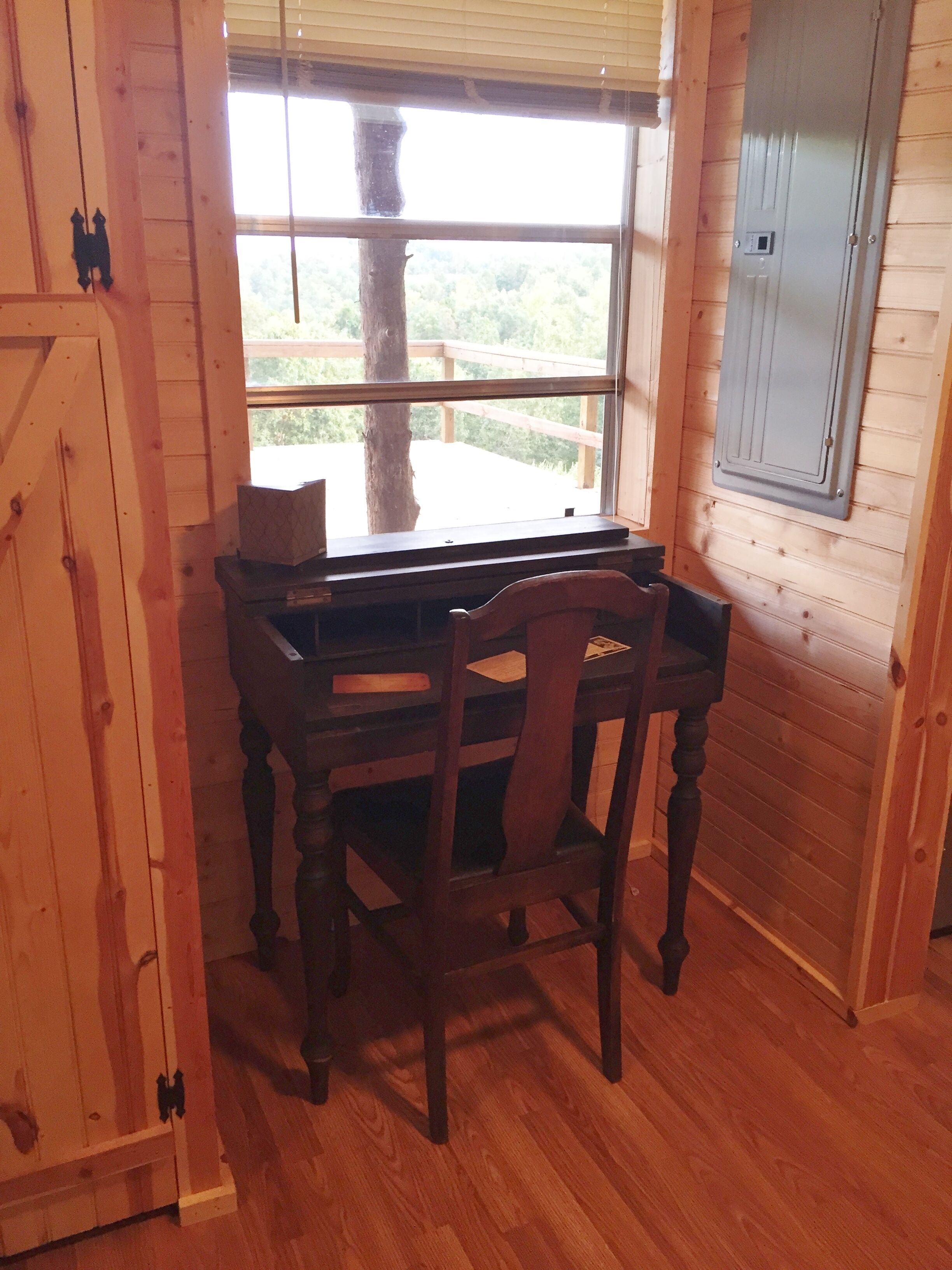 Un bureau en bois avec une chaise, dans une pièce aux murs en bois et dotée d’une fenêtre donnant sur un paysage verdoyant.