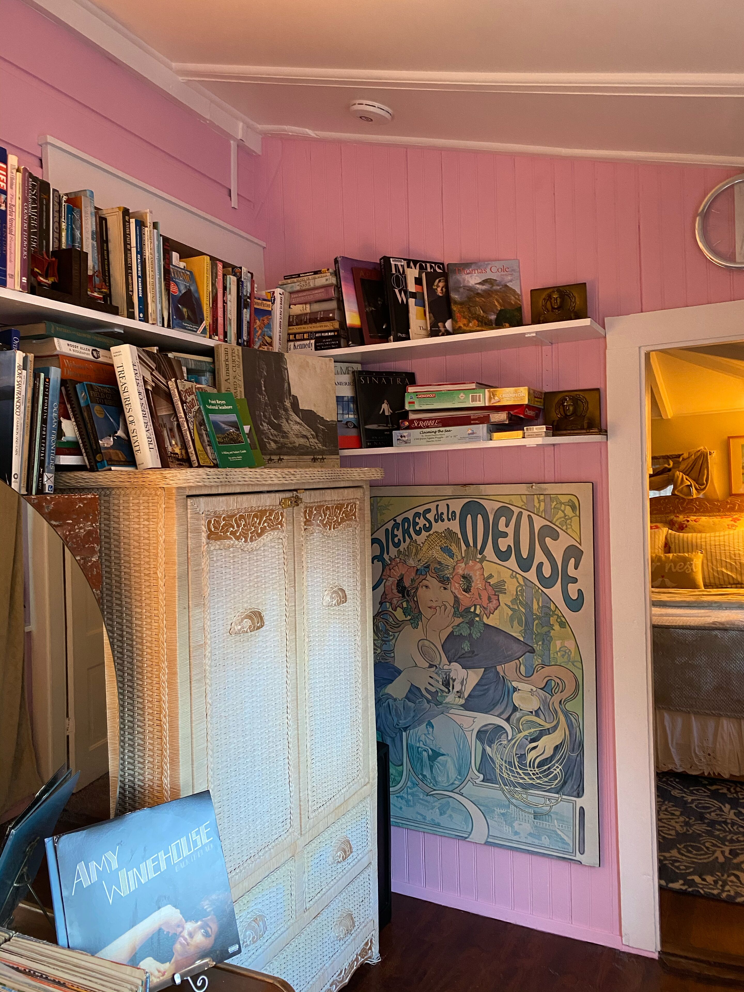 Quarto com paredes cor-de-rosa, uma estante cheia de livros, uma mala antiga e um pôster de 'Rires de la Meuse'.