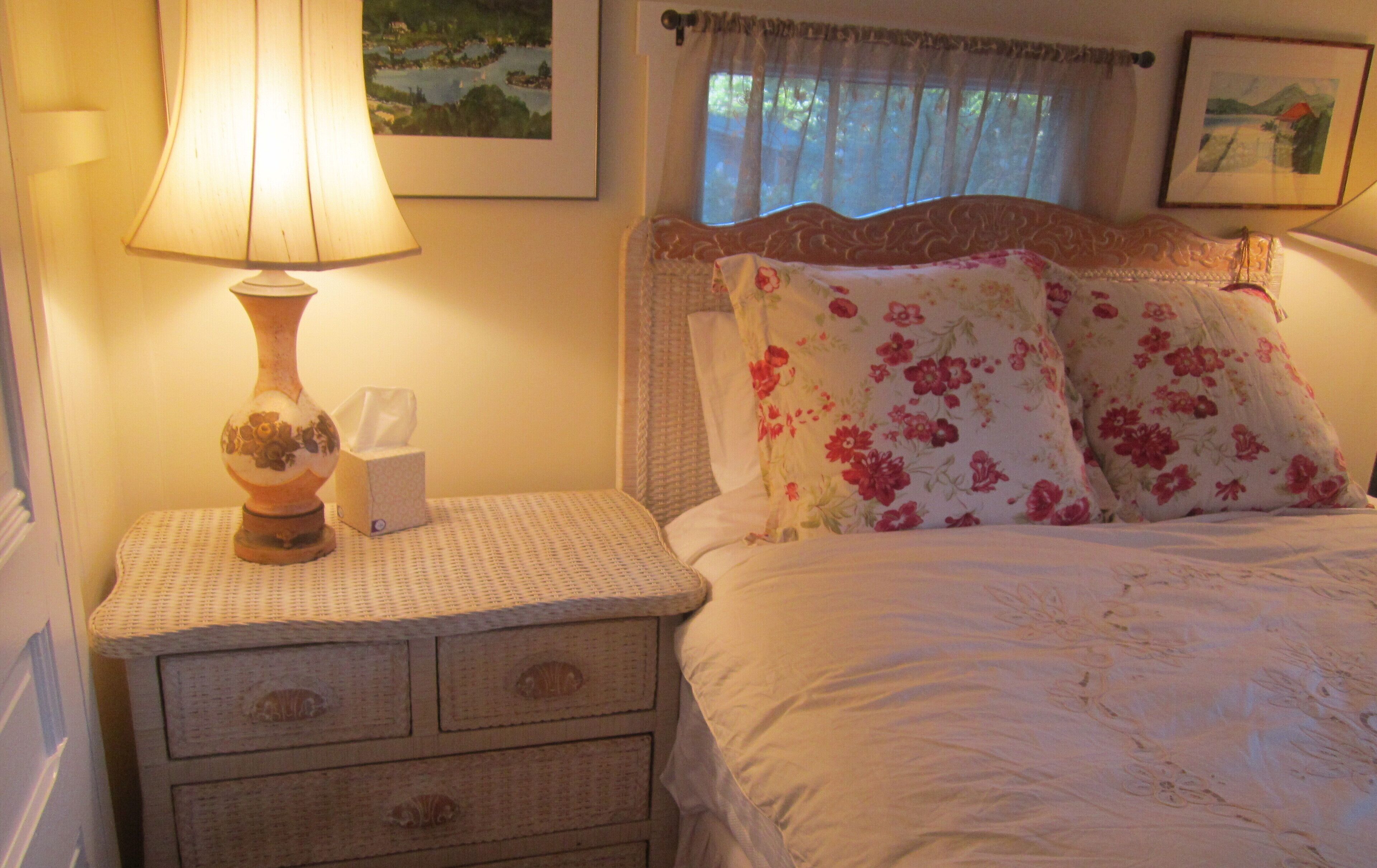 A neatly made bed with floral pillows, a bedside lamp, and a small bedside table with a tissue box.