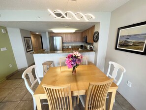 A dining area with a wooden table and chairs, a vase of flowers, and a kitchen in the background.