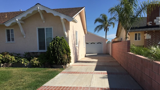 A residential house with a paved driveway, a brick wall, and a palm tree in the background.