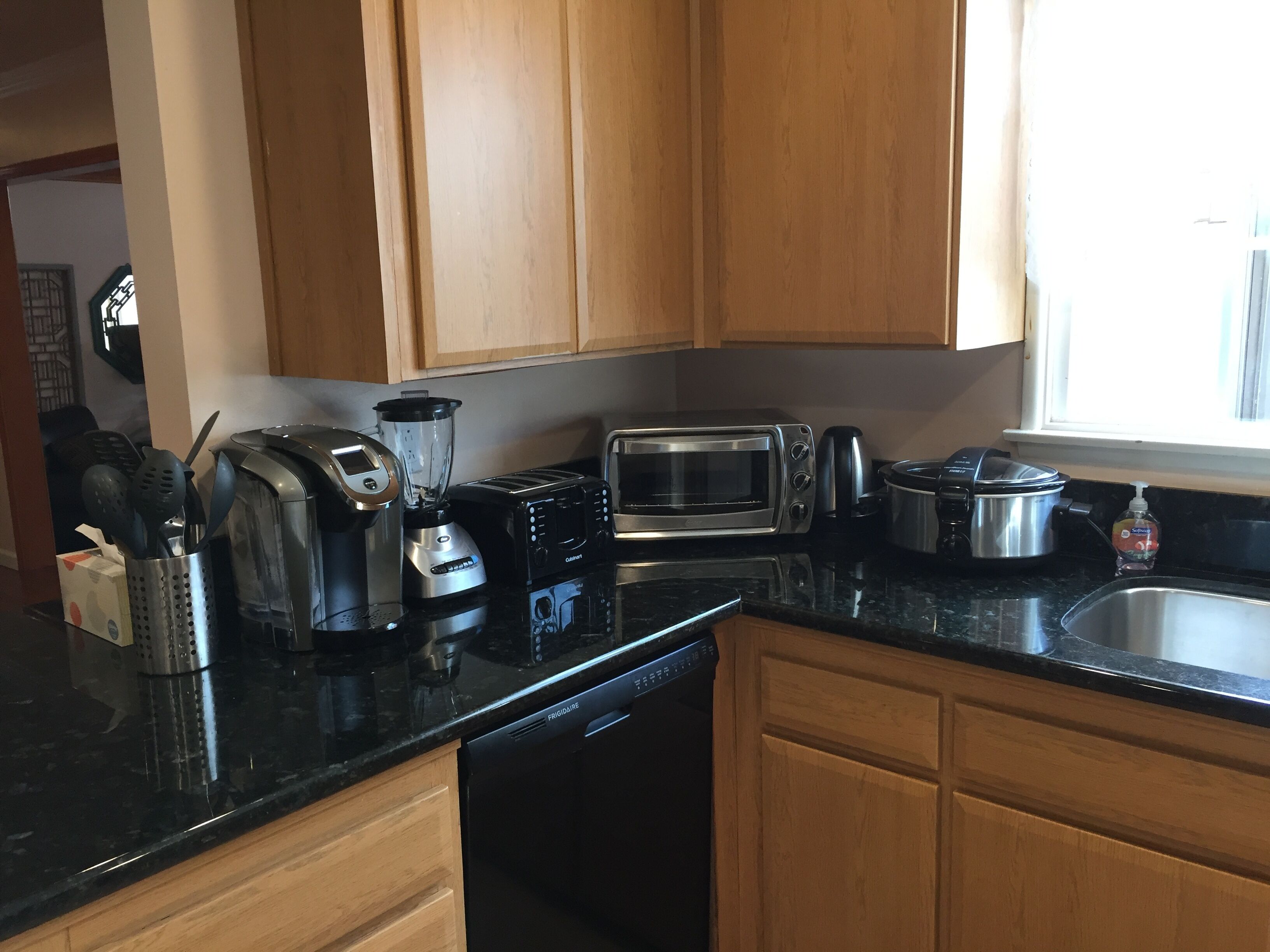 Una cocina con armarios de madera, encimera negra, cafetera, tostadora, licuadora, lavavajillas y un lavabo.