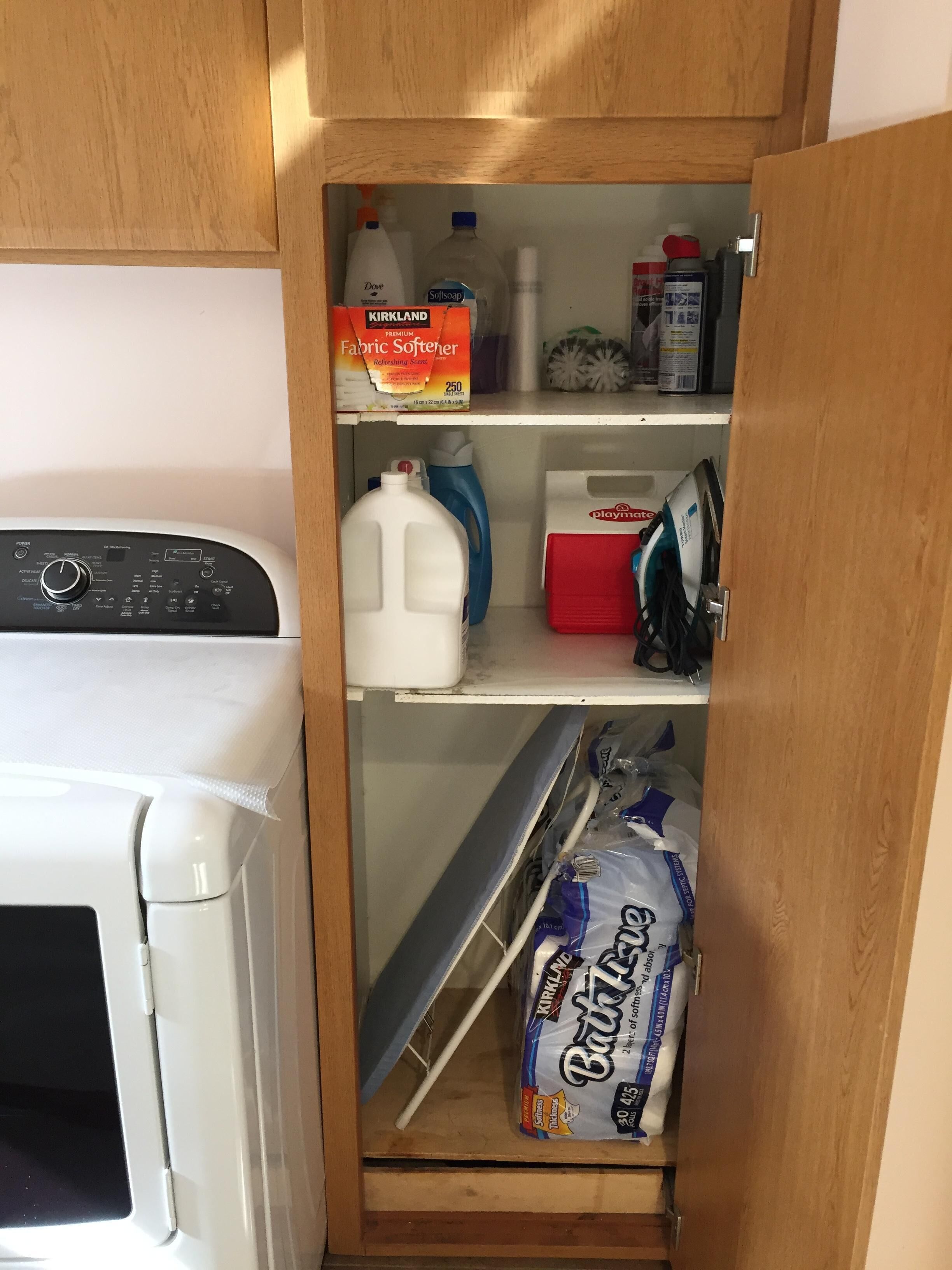 A laundry room with a washing machine, a shelf with Kirkland fabric softener, a bottle of Dove soap, a bottle of Softsoap, a Playmate iron, and a bag of Kirkland detergent.