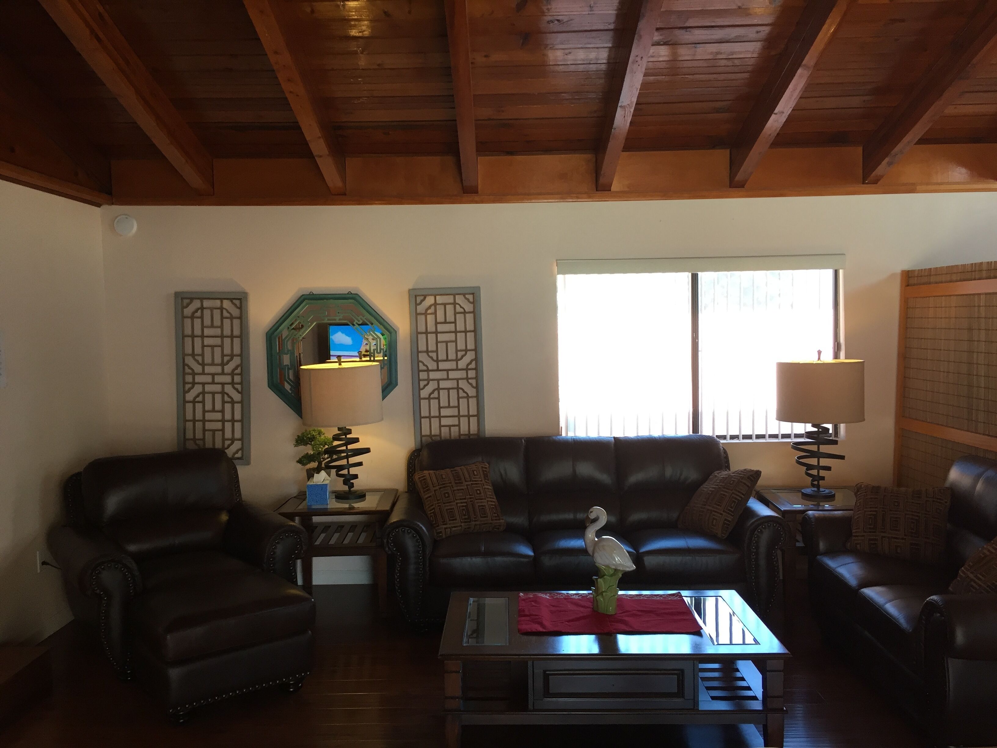 A living room with a wooden ceiling, a black leather sofa, a coffee table with a vase, and a window with blinds.