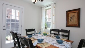 Een eetkamer met een houten tafel, zwarte stoelen en een kroonluchter. Er is een raam met witte gordijnen en een ingelijst schilderij aan de muur.
