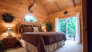 A wooden cabin bedroom with a large bed, a nightstand with a lamp, and a window overlooking a deck. - Two Master Suites, Great for 2 couples, Walking distance to Thunder Ridge Arena (Ridgedale)