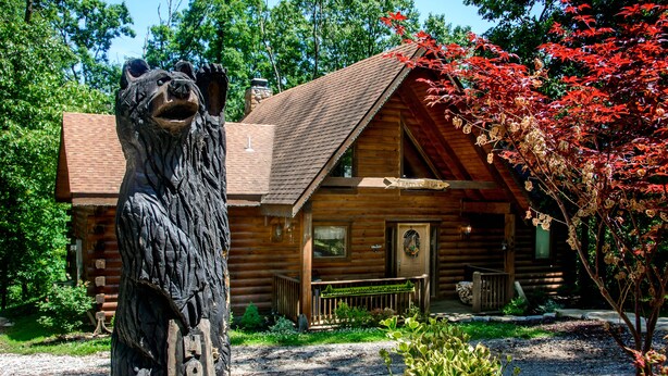 A wooden cabin with a porch, surrounded by trees and a bear sculpture. - Two Master Suites, Great for 2 couples, Walking distance to Thunder Ridge Arena (Ridgedale)