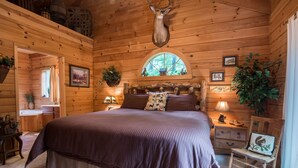 A log cabin bedroom with a large bed, wooden walls, a deer head mounted on the ceiling, a window with a view of trees, and a small desk with a lamp. - Two Master Suites, Great for 2 couples, Walking distance to Thunder Ridge Arena (Ridgedale)
