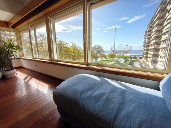 A room with a large window, wooden flooring, a potted plant, and a view of a building and a street.