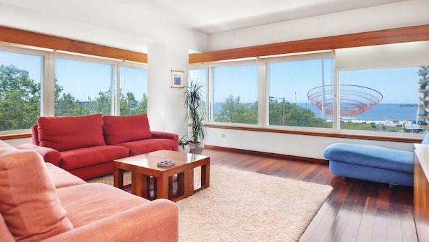 Un salon moderne avec un canapé rouge, un fauteuil bleu et une table basse en bois. De grandes fenêtres offrent une vue sur la mer et sur une structure rouge.