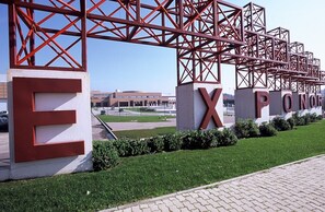 Entrada de edifício com letras vermelhas grandes formando o nome, rodeada por estrutura metálica e vegetação.