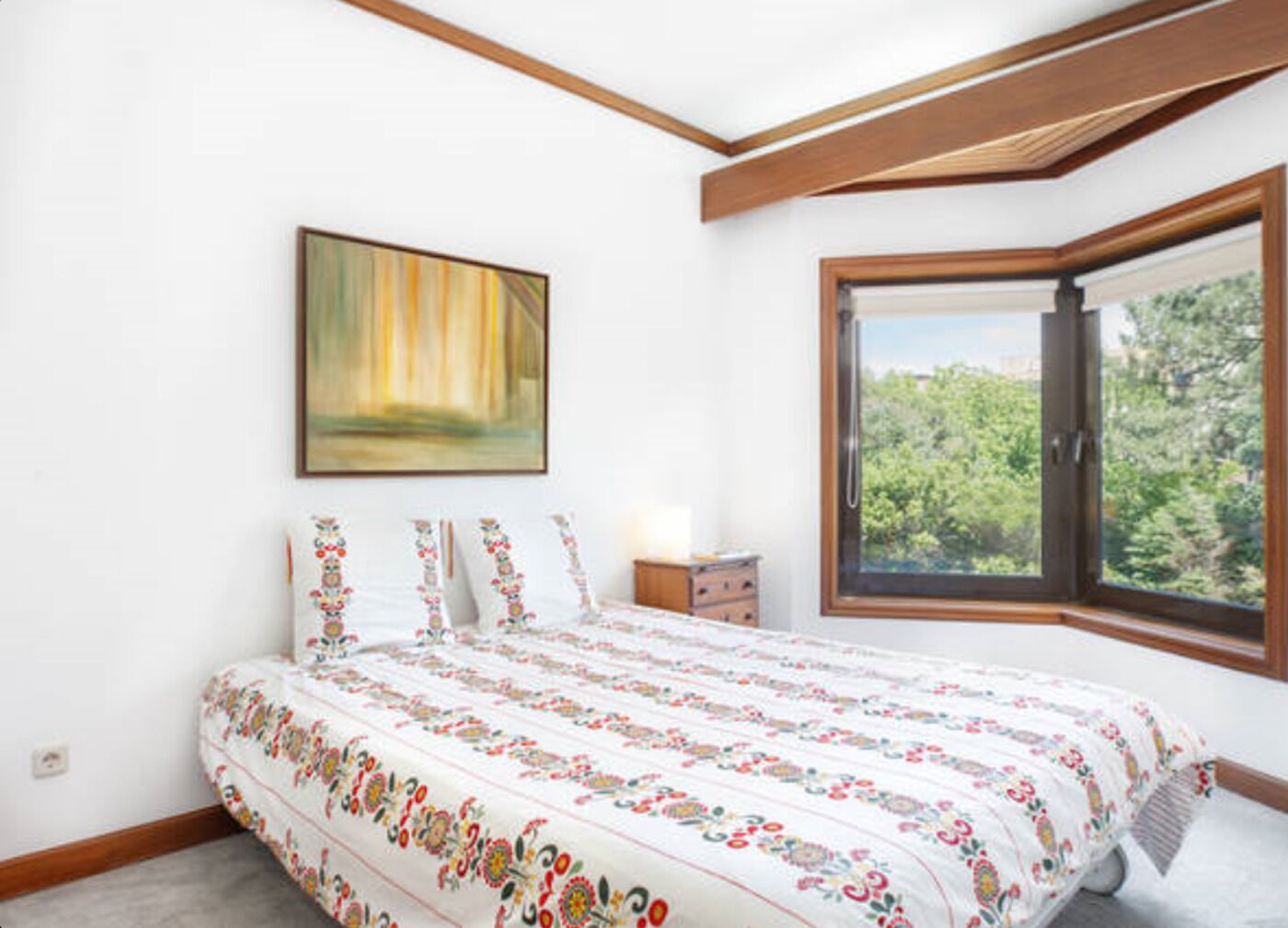 A bedroom with a large bed, a nightstand, a painting on the wall, and a window with a view of trees.