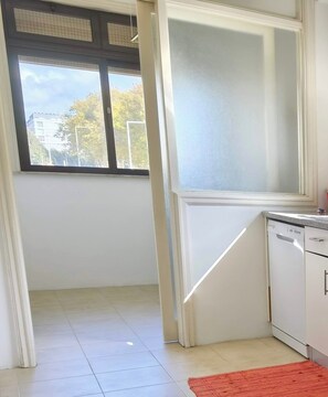 A kitchen with a dishwasher, a window with blinds, and a tiled floor.