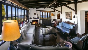 A cozy living room with a fireplace, a sofa, a coffee table, and a view of the outdoors through large windows. - Ampelos Estate (Aigialeia)