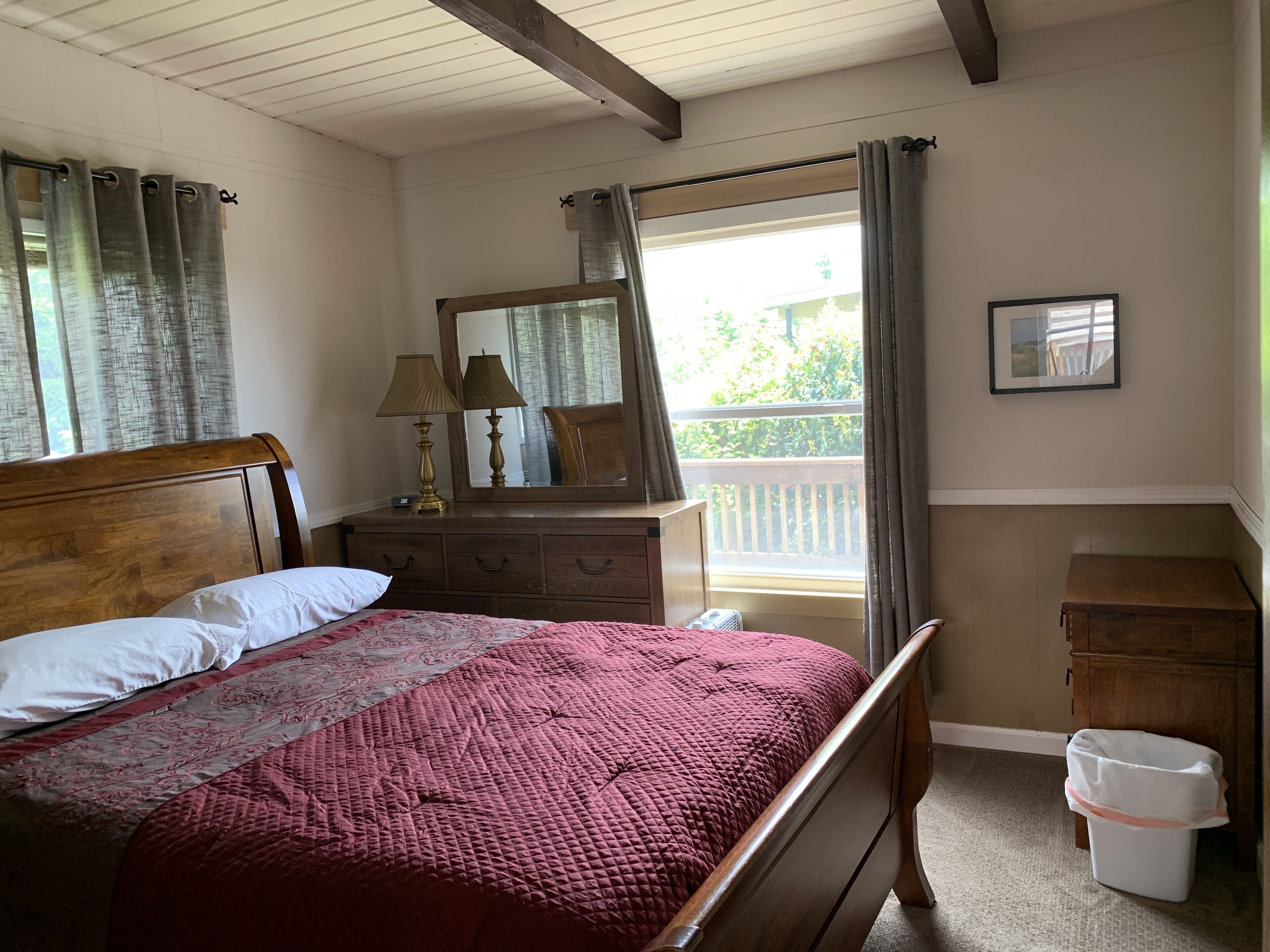 Un dormitorio con cama de madera, cómoda, espejo, ventana con cortinas y un pequeño cubo de basura.
