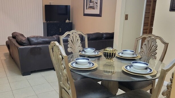 A dining area with a round glass table set for four, surrounded by ornate chairs. A dark leather sofa and a cabinet with a television are in the background. - Cozy Condo For Rent In Melbourne Florida.  5 Miles from the beach (Melbourne)