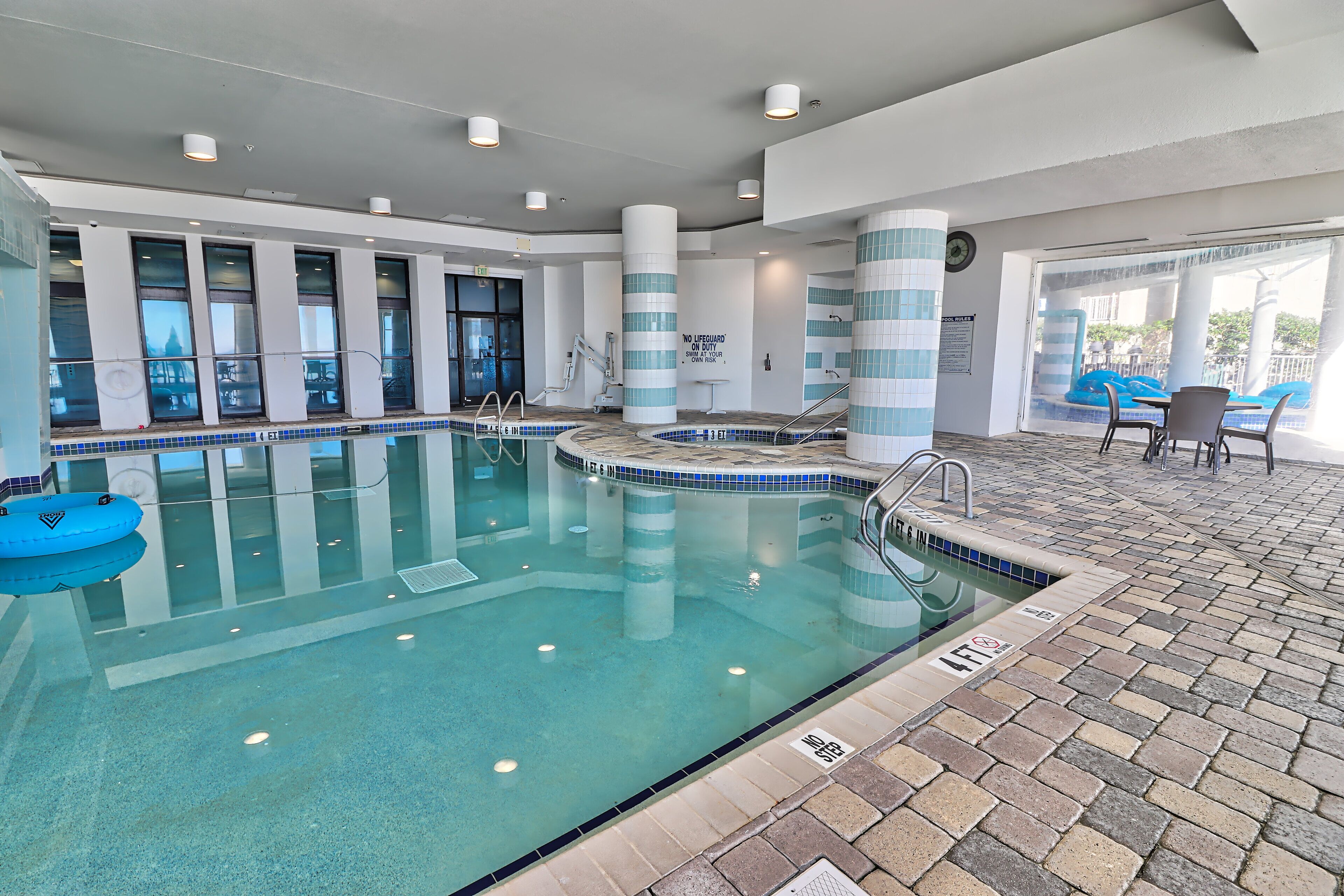 Piscina interna com água cristalina, posto de salva-vidas e área com cadeiras e mesa à beira da piscina.