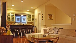 Una sala acogedora con una mesa de centro de vidrio, un sofá y un área de cocina con taburetes de bar.