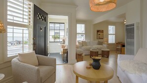 Ein geräumiges Wohnzimmer mit einer dunklen Tür, weißen Wänden und einem großen Fenster. Es steht ein runder Holztisch mit einer Vase, ein weißes Sofa und ein Sessel.