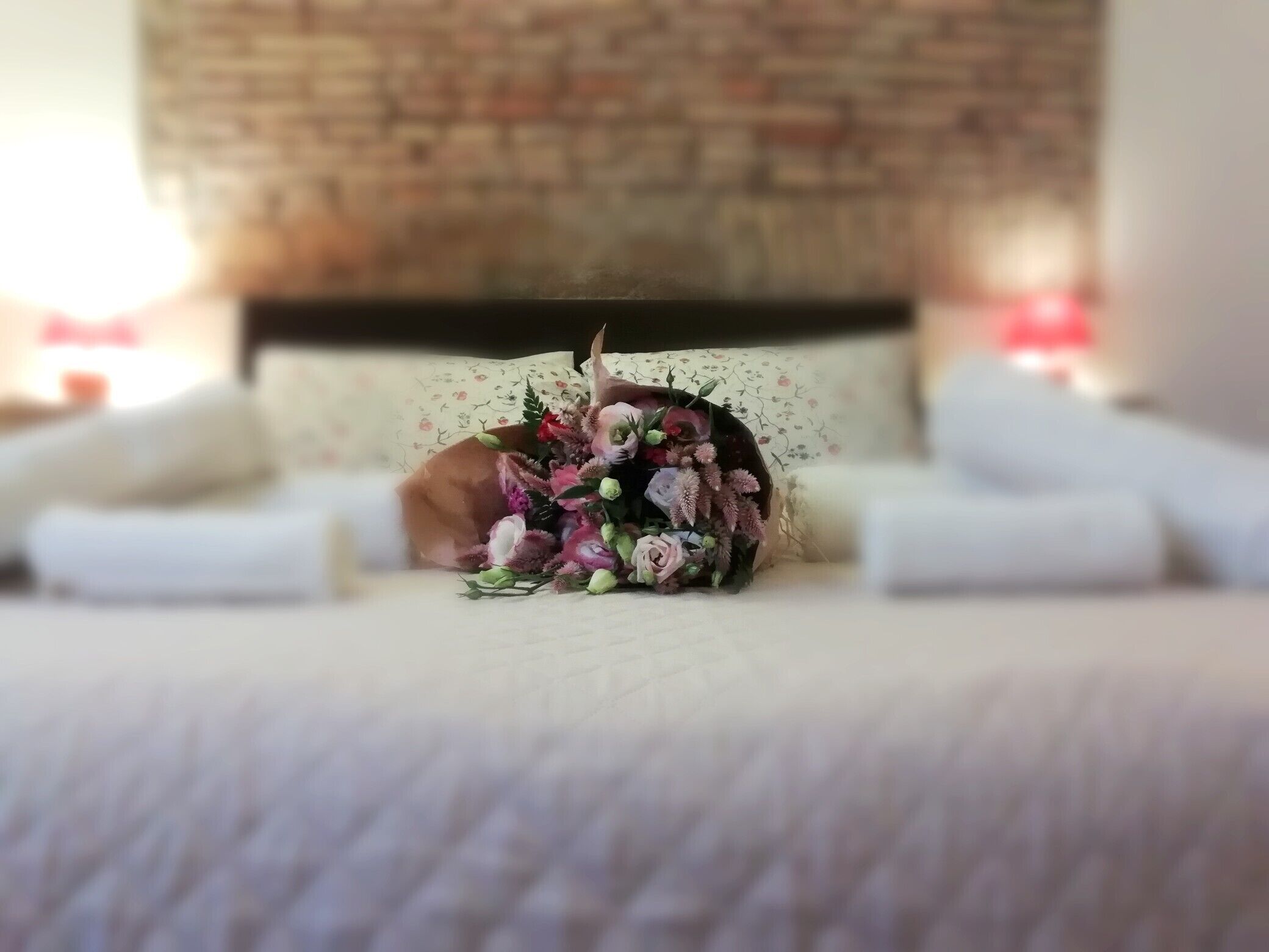 A neatly made bed with a floral arrangement in the center, a brick wall behind, and a floral-patterned pillow.